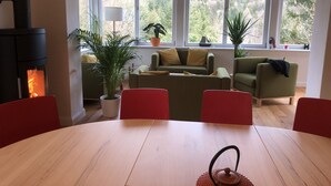 Uma sala de estar aconchegante com lareira, sofás verdes, uma mesa de jantar e janelas amplas com vista para árvores.