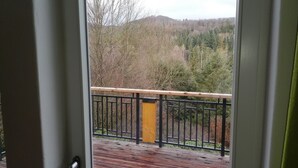 Um deck de madeira com guarda-corpo preto e um poste amarelo, com vista para uma área arborizada com árvores e colinas ao longe.