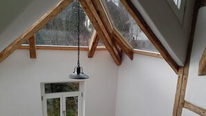 Quarto com teto inclinado, luminária pendente, janela com moldura branca e vigas de madeira.