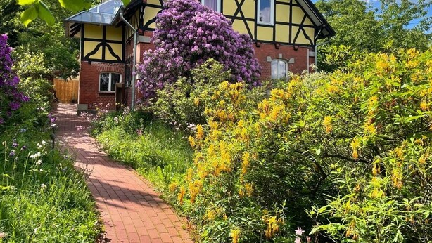 Uma casa tradicional em estilo de enxaimel, com um caminho de tijolos que leva à entrada, cercada por vegetação exuberante e plantas floridas.