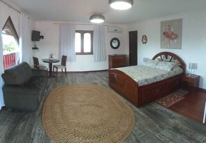 A bedroom with a bed, a round woven rug, a wooden headboard, a nightstand, a bedside table, a wall-mounted TV, a window with curtains, a clock, and a door. - Paracuru Kitefriends Pousada (Paracuru)