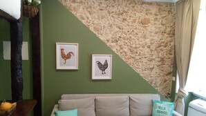 A room with a green wall, a stone accent wall, a beige sofa with a teal pillow, and two framed rooster pictures.