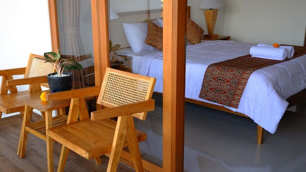 A hotel room with a wooden bed, white bedding, a patterned throw, a wooden chair with a potted plant, and a glass partition. - Bale Delod Guest House (Ubud)