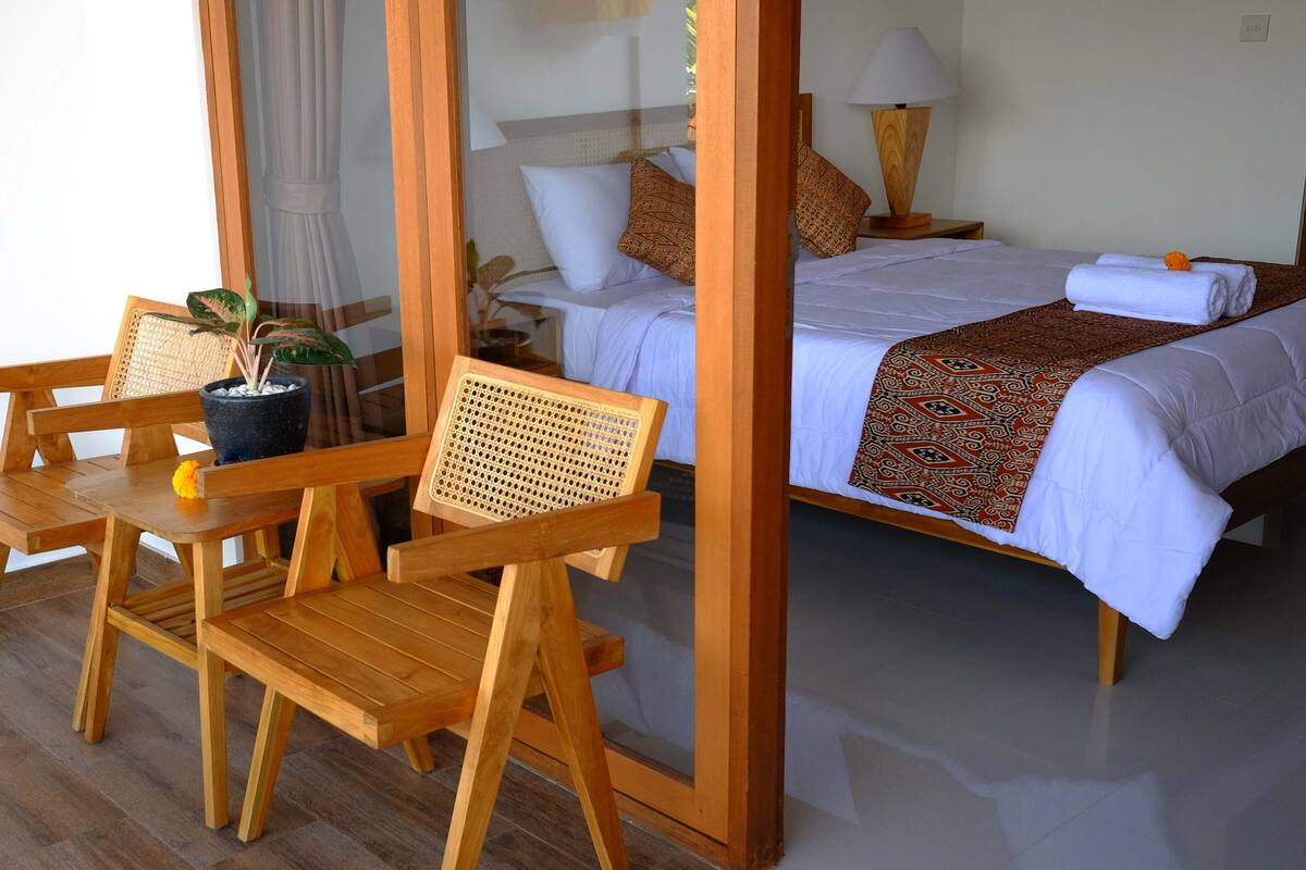 A hotel room with a wooden bed, white bedding, a patterned throw, a wooden chair with a potted plant, and a glass partition.