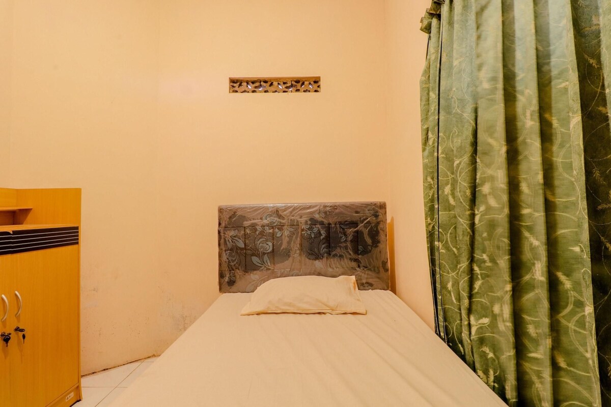 A small, single-bed room with a wooden headboard, a small shelf unit, and a green curtain.