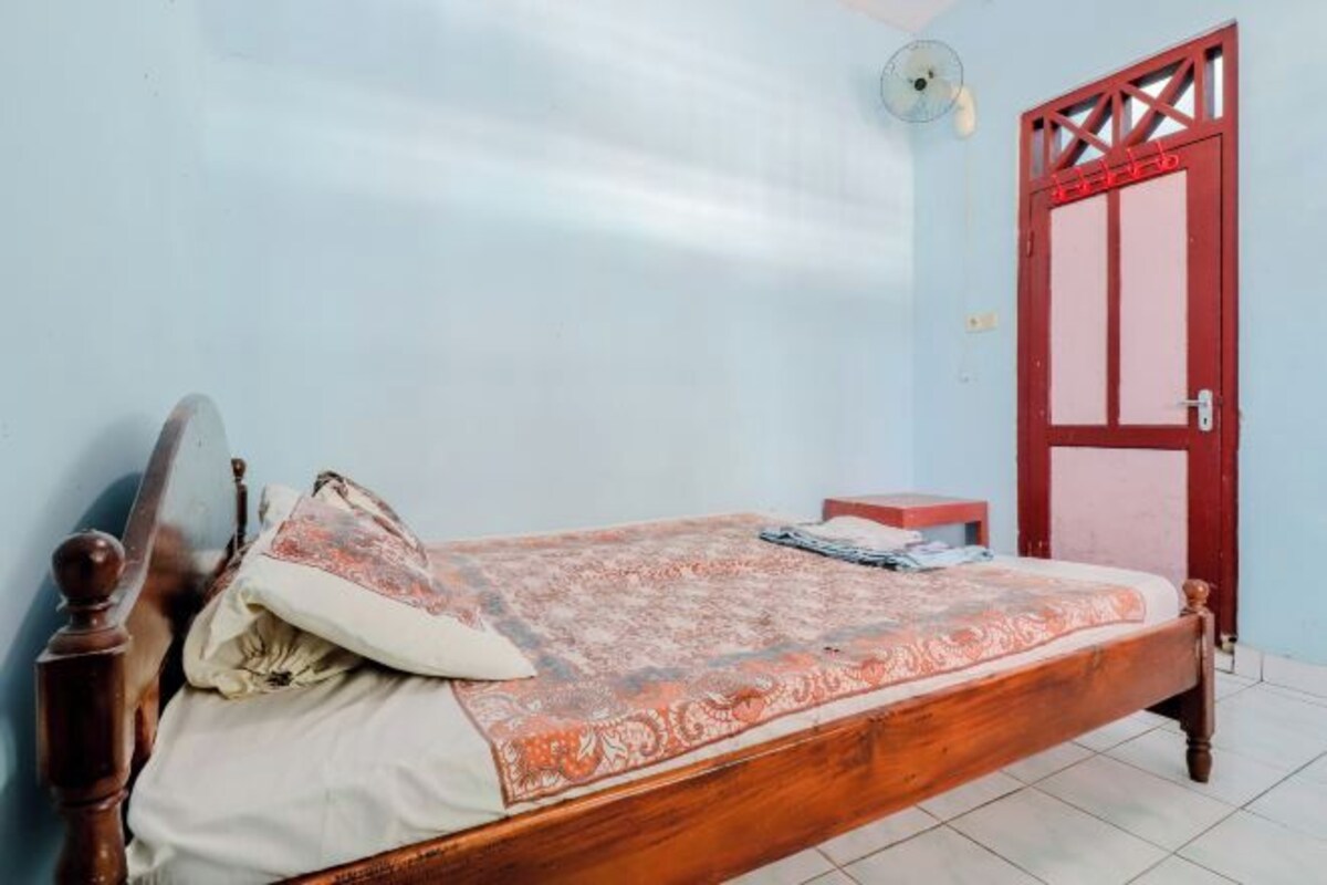 A single bed with a wooden frame and a patterned bedspread, a small bedside table, and a red door with a window.