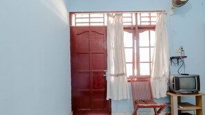 A room with a wooden door, a window with white curtains, a wooden chair, and a small wooden TV stand with an old television. - Losmen Parikesit (Parangtritis)