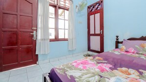 A bedroom with a bed, a window with curtains, a door, and a fan on the wall. - Losmen Parikesit (Parangtritis)