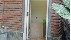 A bathroom with a brick wall, a tiled floor, and a green trash bin. - Majapahit Homestay by Bhagaskara (Bejijong)