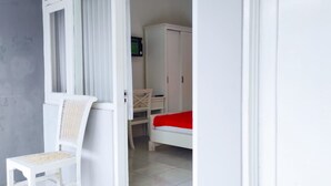 A room with a white chair, a bed with a red blanket, a desk, and a wardrobe. - Ratu Guesthouse (Depok)