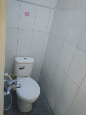 A small, compact bathroom with a white toilet, a visible pipe, and a tiled floor. - Ratu Guesthouse (Depok)