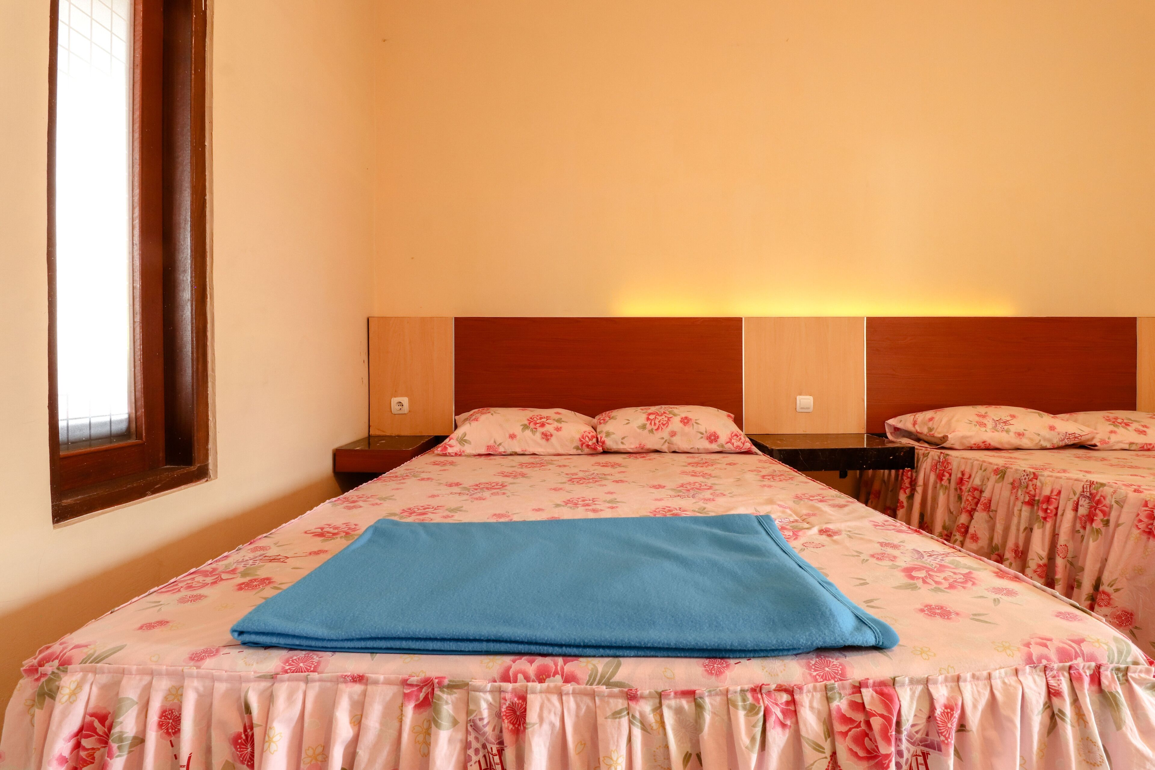 Quarto de hotel com duas camas, cada uma com roupa de cama floral e uma manta azul em uma delas. Há mesas de cabeceira com abajures e uma janela com persianas.