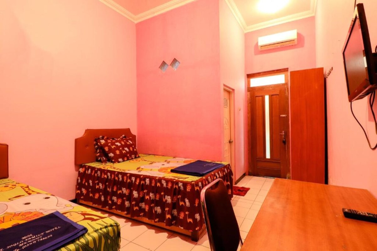 A bedroom with pink walls, a bed with a colorful bedspread, a wooden table, a chair, and a TV mounted on the wall.
