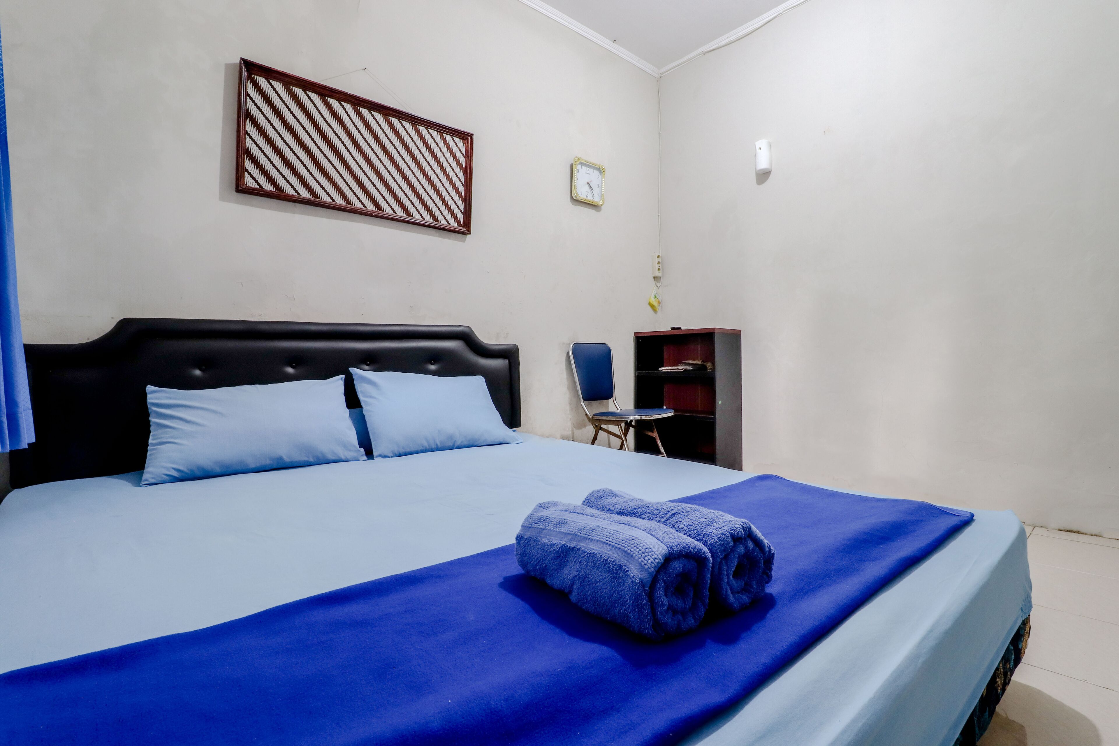 Una cama bien hecha con una colcha azul y almohadas, un cabecero de madera, un escritorio pequeño con una silla y un reloj fijado a la pared.
