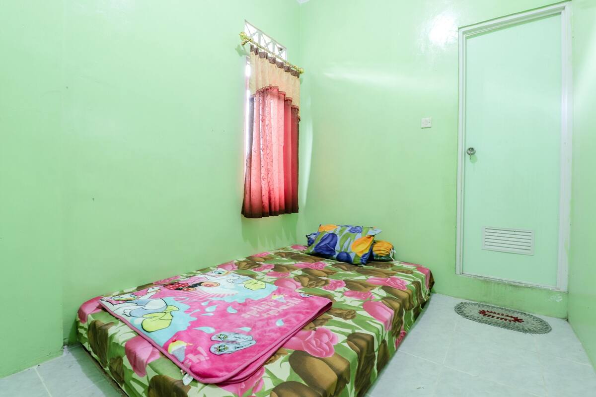 A small, single-bed room with a floral bedspread, a window with red curtains, and a door with a vent.