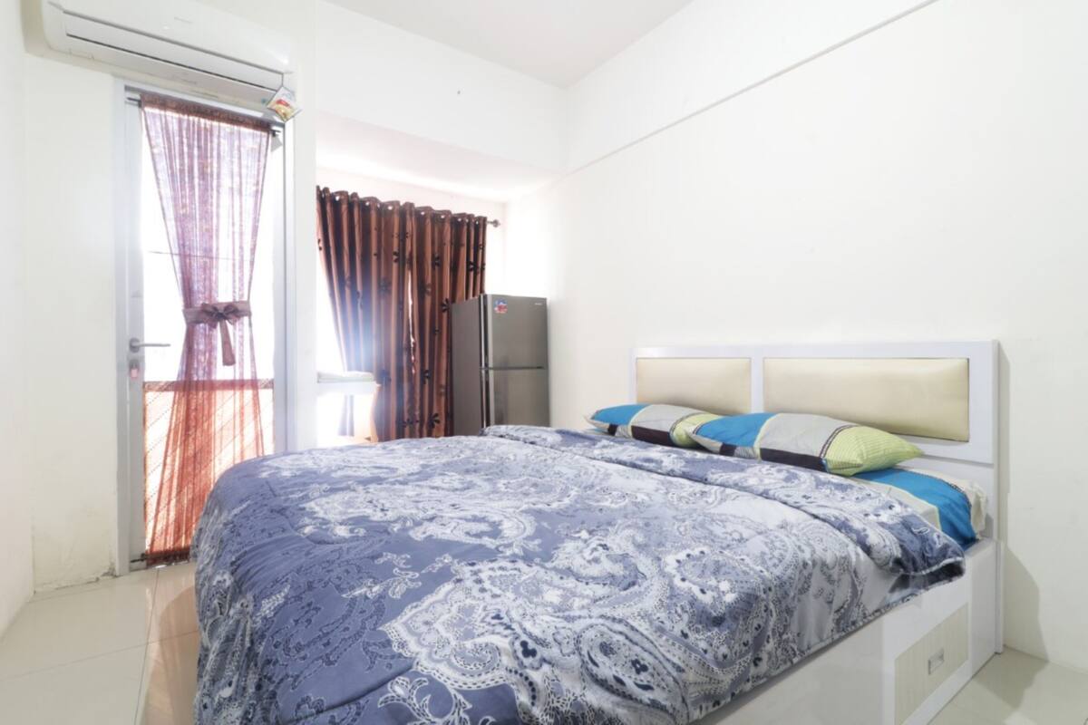 A bedroom with a bed, curtains, a refrigerator, and a window.