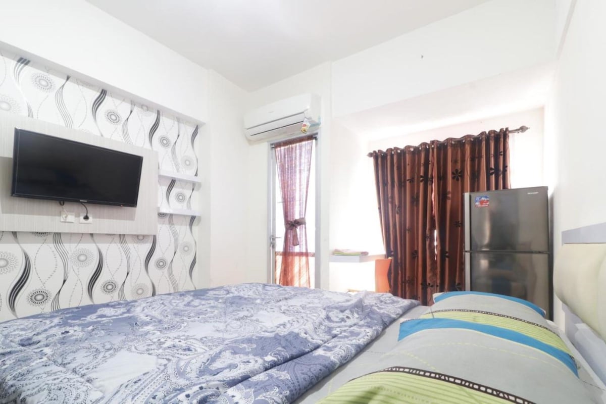 Une chambre à coucher comprenant un lit, un téléviseur fixé au mur, un appareil de climatisation, un réfrigérateur et une fenêtre avec des rideaux.
