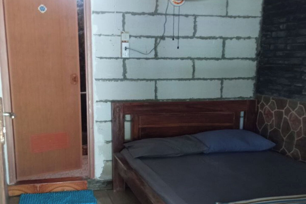 A bedroom with a wooden bed, a blue mattress, a wooden headboard, a wall-mounted fan, and a tiled floor.