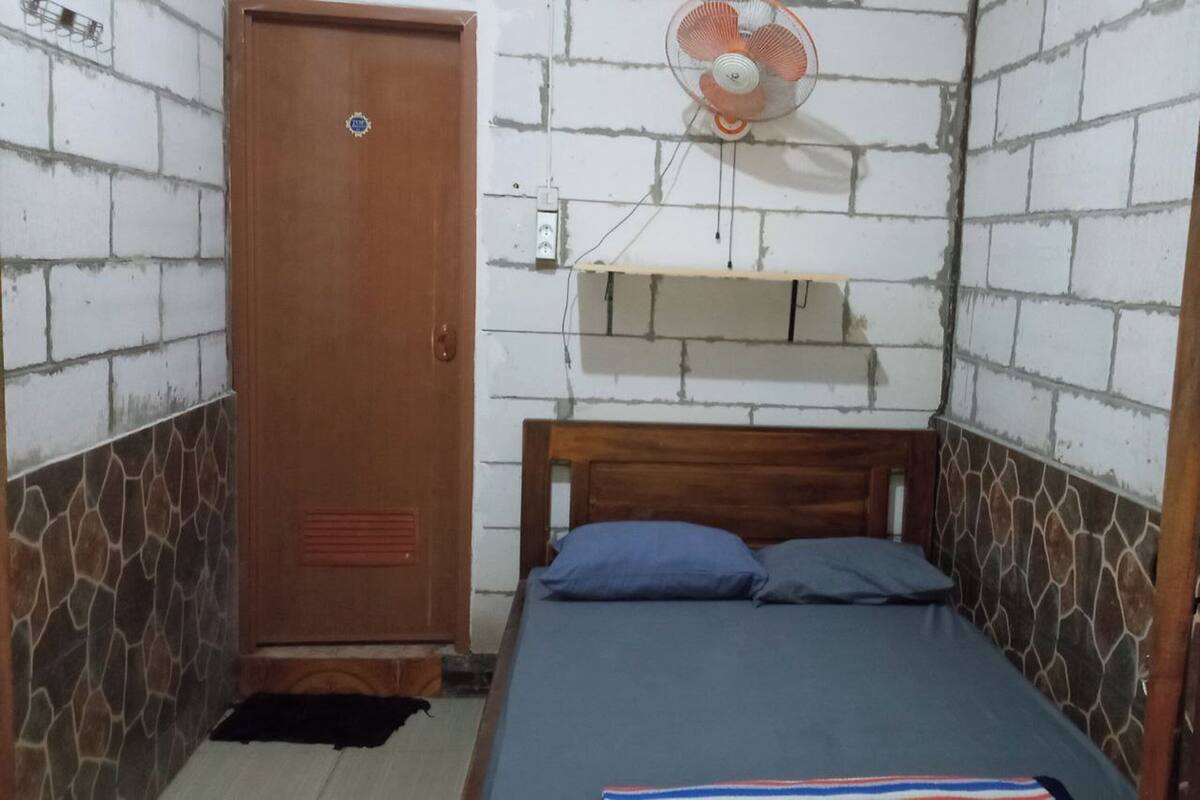 A small, single-bed room with a wooden headboard, a wooden wardrobe, a wall-mounted fan, and a small shelf.