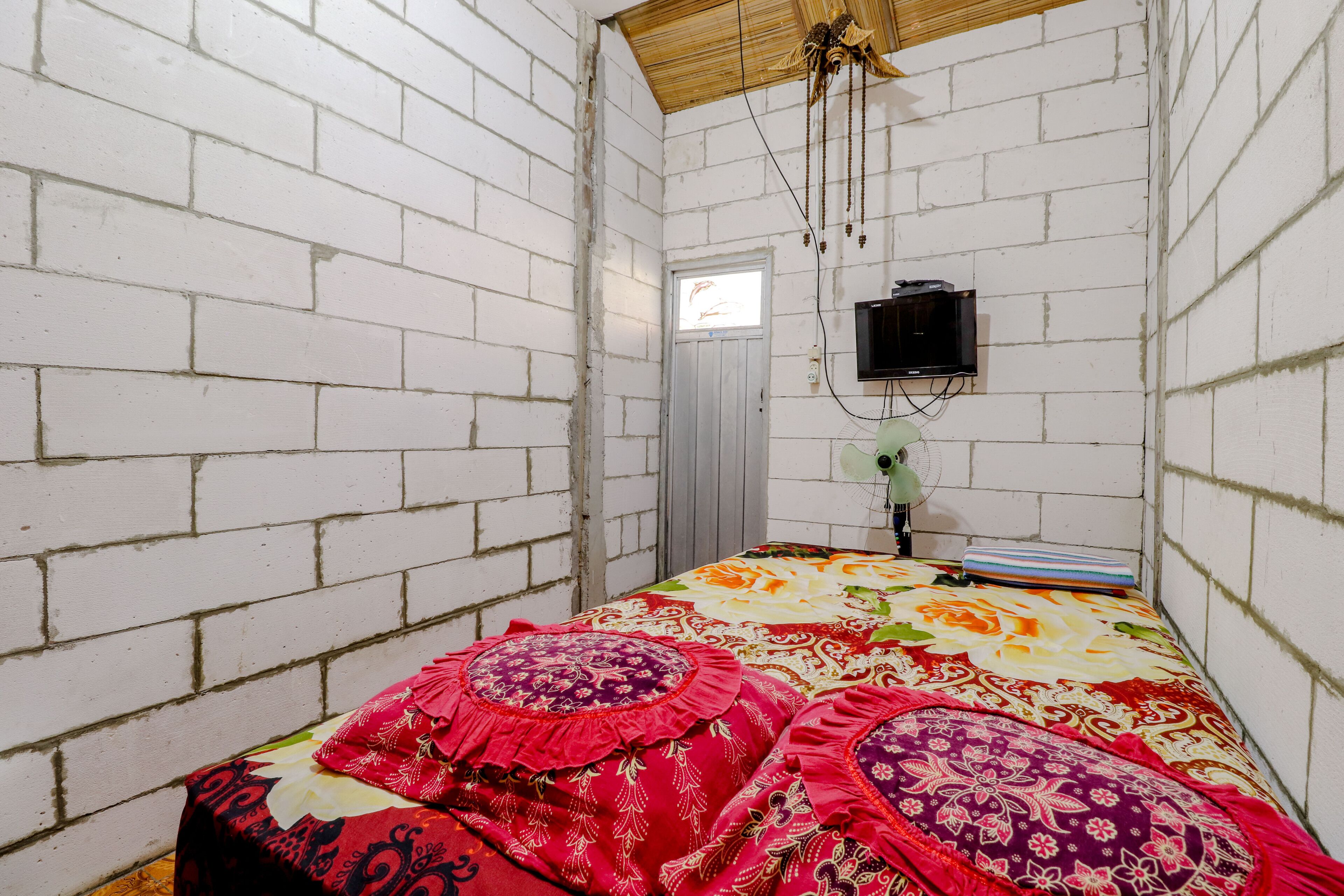 A small, narrow room with white brick walls, a single bed with colorful bedding, a small window, and a television mounted on the wall.