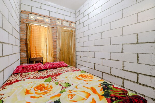 Una habitación pequeña y estrecha con paredes de ladrillo blanco, una puerta de madera con espejo y una cama con colcha estampada de flores.