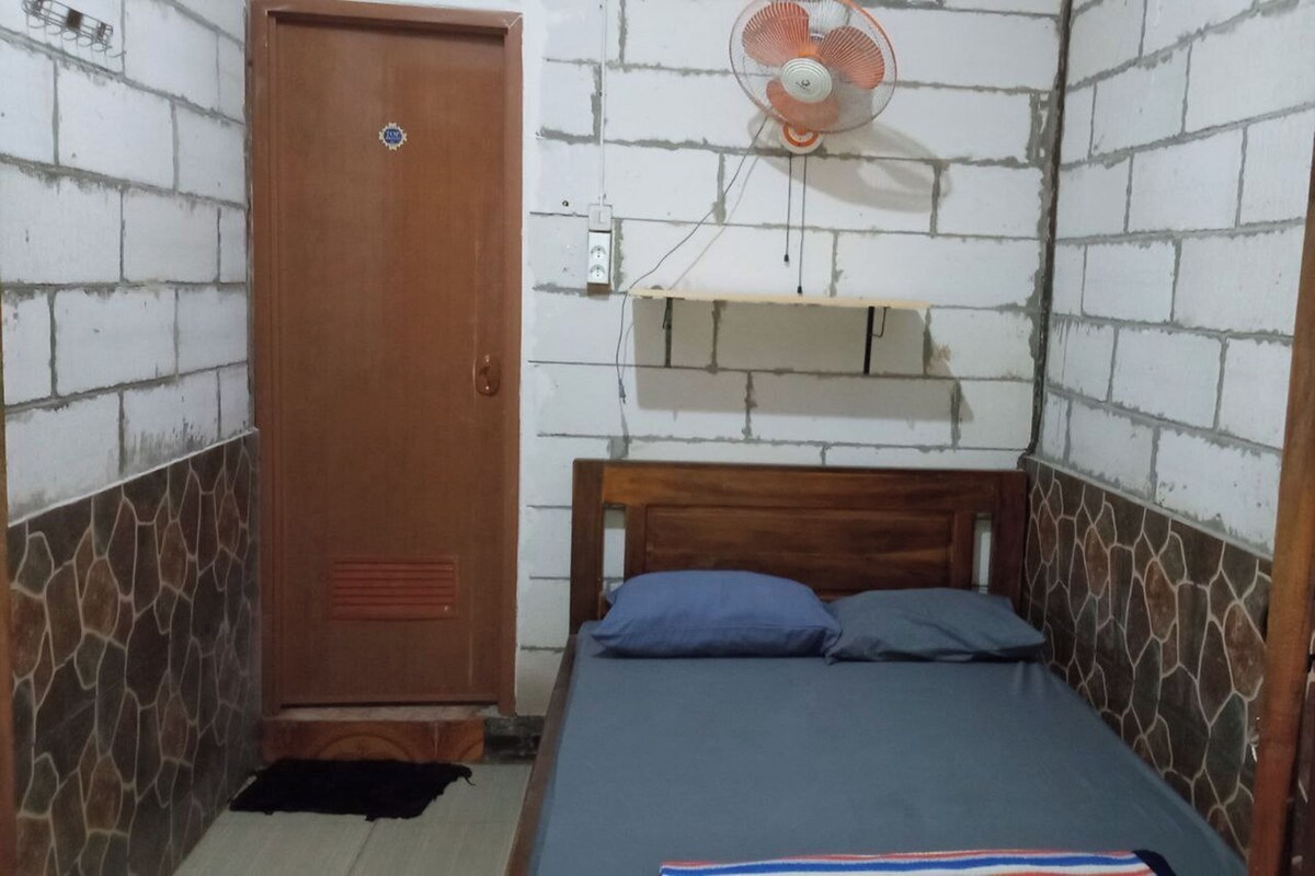 A small, single-bed room with a wooden headboard, a wooden wardrobe, a wall-mounted fan, and a small shelf.
