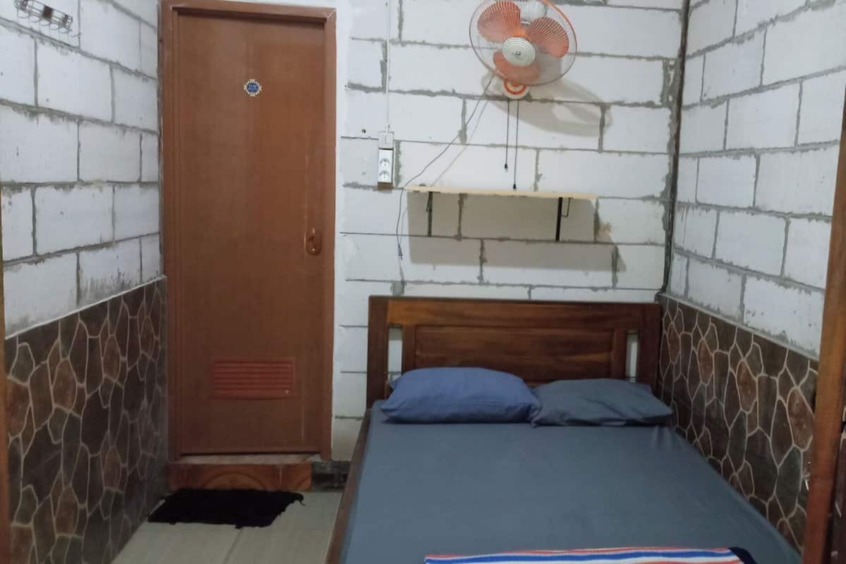 A small, single-bed room with a wooden headboard, a wooden wardrobe, a wall-mounted fan, and a small shelf.