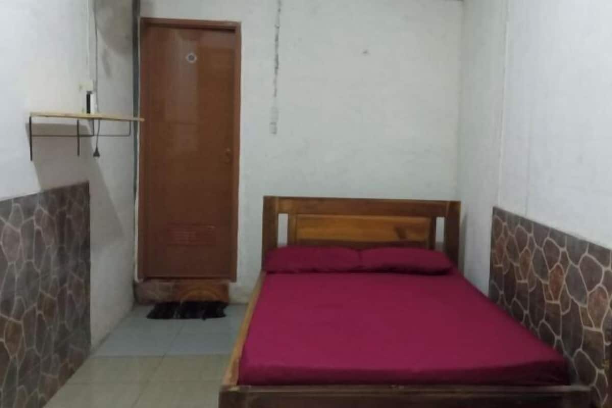 A small, single-bed room with a wooden headboard, a red bedspread, a wardrobe, and a stone-patterned wall.