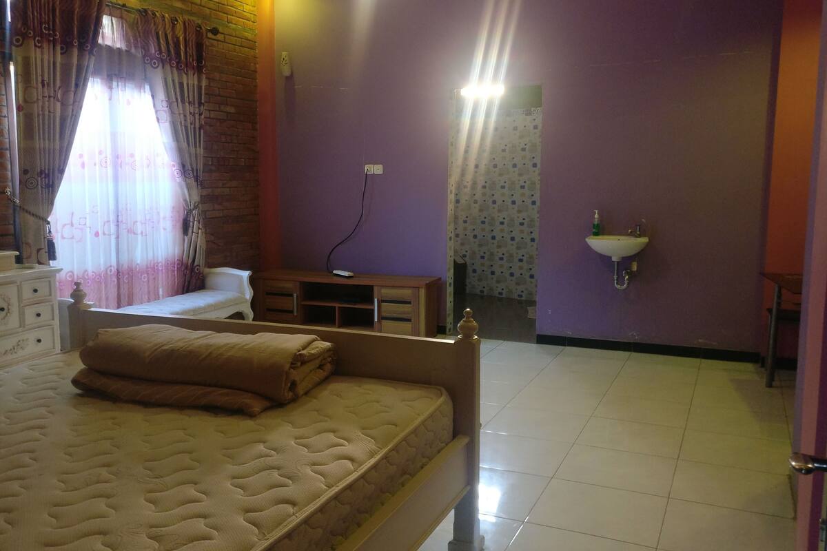 Un dormitorio con cama, mesita de noche, silla, ventana con cortinas, lavamanos empotrado en la pared y pared de ladrillo.