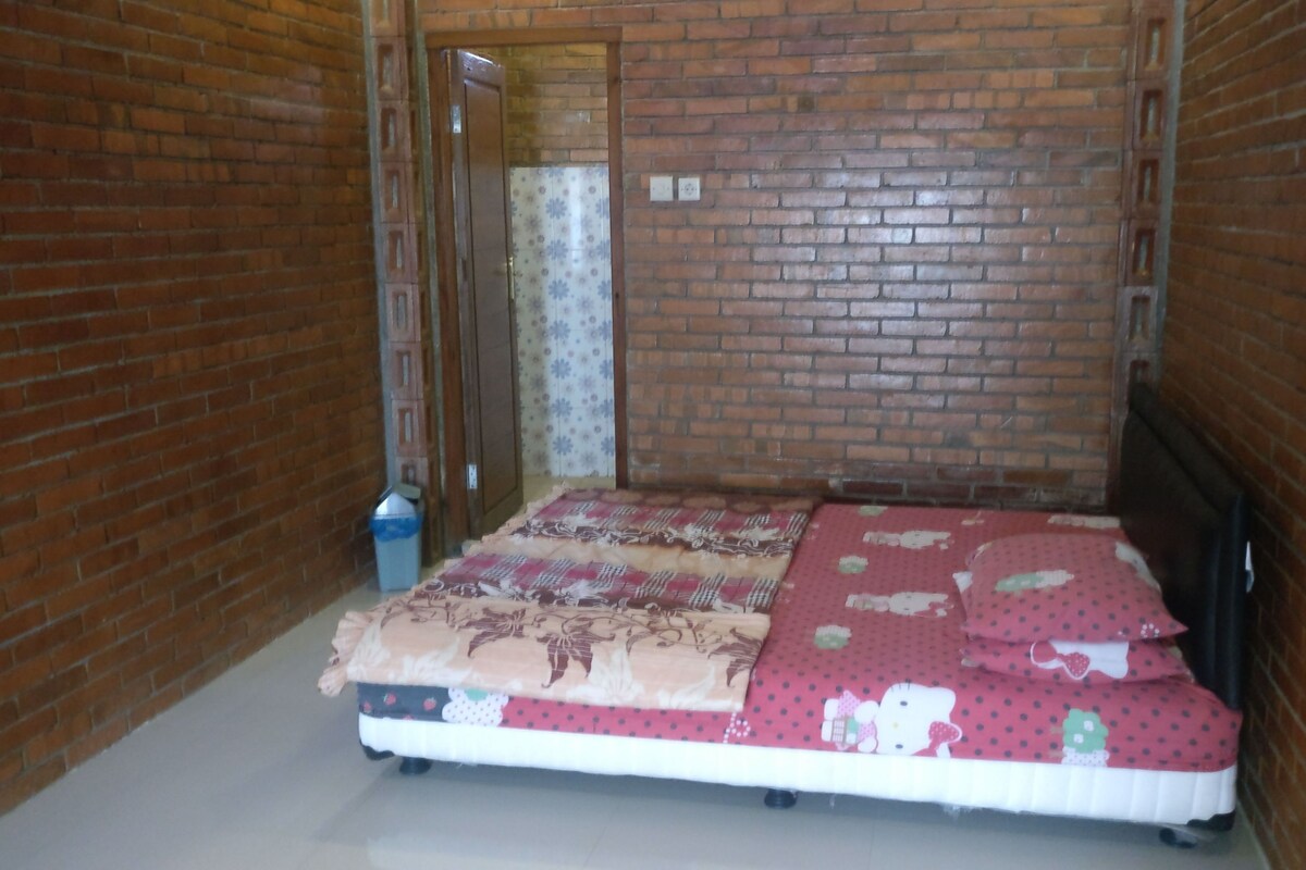A narrow room with a bed, a trash bin, and a door leading to another room.