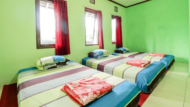 Una habitación con cuatro camas, cada una con una colcha y almohadas de diferente color. Hay ventanas con cortinas rojas y un equipo de aire acondicionado en la pared.