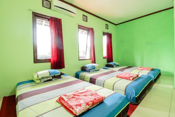 A room with four beds, each with a different color bedspread and pillows. There are windows with red curtains, and an air conditioning unit on the wall. - Penginapan Roro Mendut Syariah by Zuzu (Mungkid)