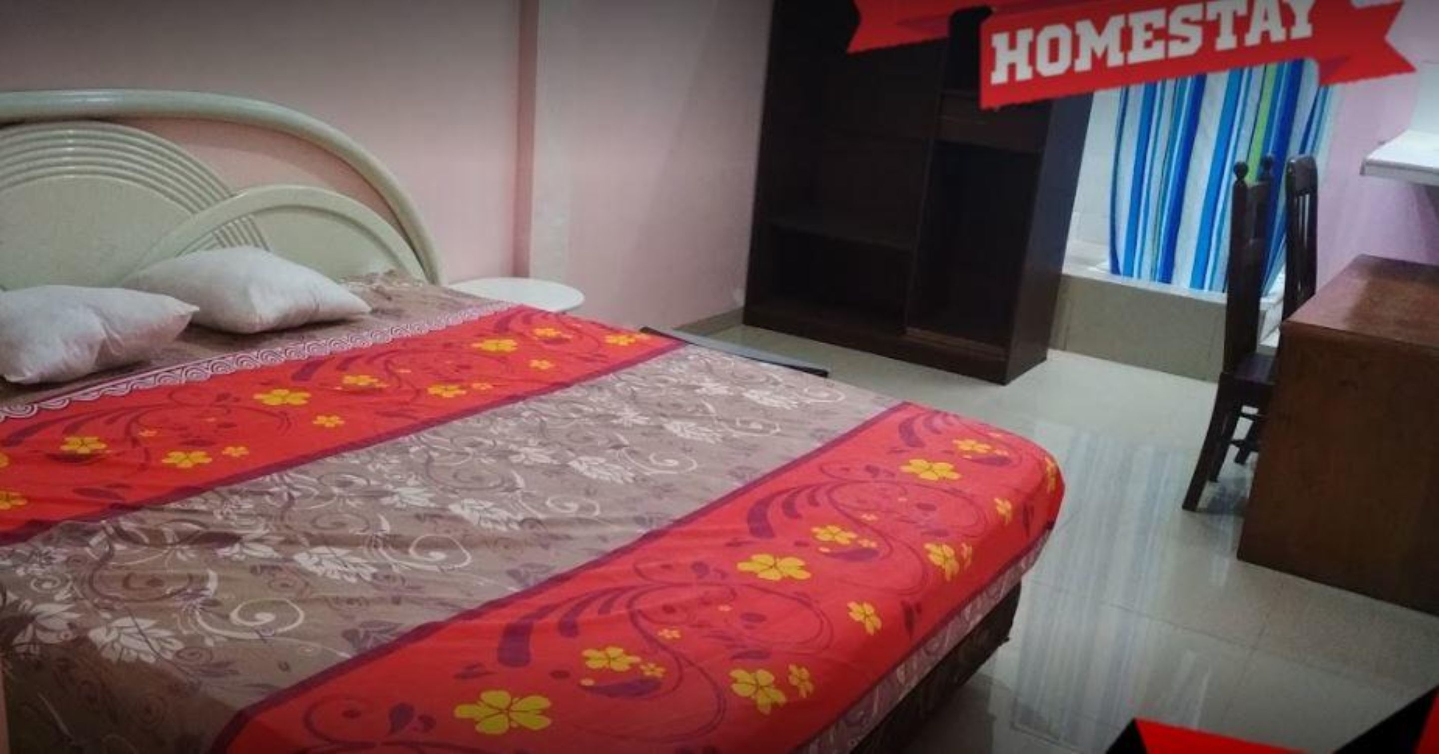 A hotel room with a bed, pillows, a red and yellow floral bedspread, a wooden headboard, a wooden nightstand, and a red sign that reads "HOMESTAY".
