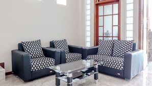 A living room with two black sofas and patterned cushions, a glass coffee table, and a large window with a wooden frame and a clock.