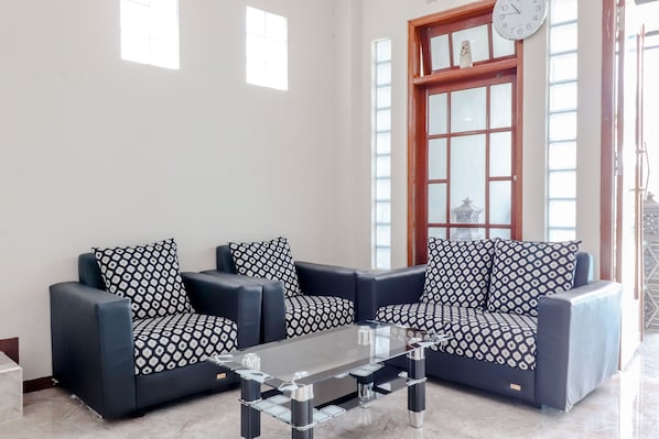 Una sala de estar con dos sofás negros y cojines estampados, una mesa de centro de vidrio y un ventanal grande con marco de madera y un reloj.