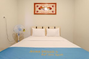Una habitación con una cama, un ventilador de pared y un cartel enmarcado sobre la cama.