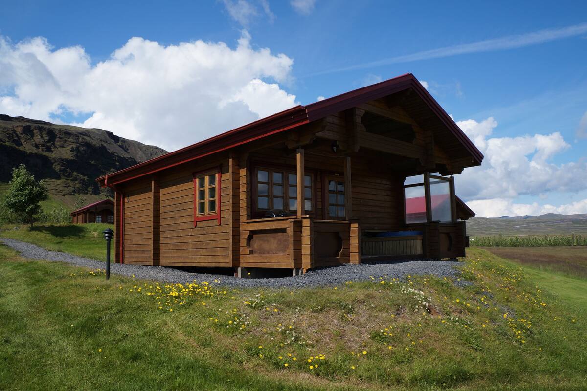 A wooden cabin with a porch, situated on a grassy hill with a mountain in the background.
