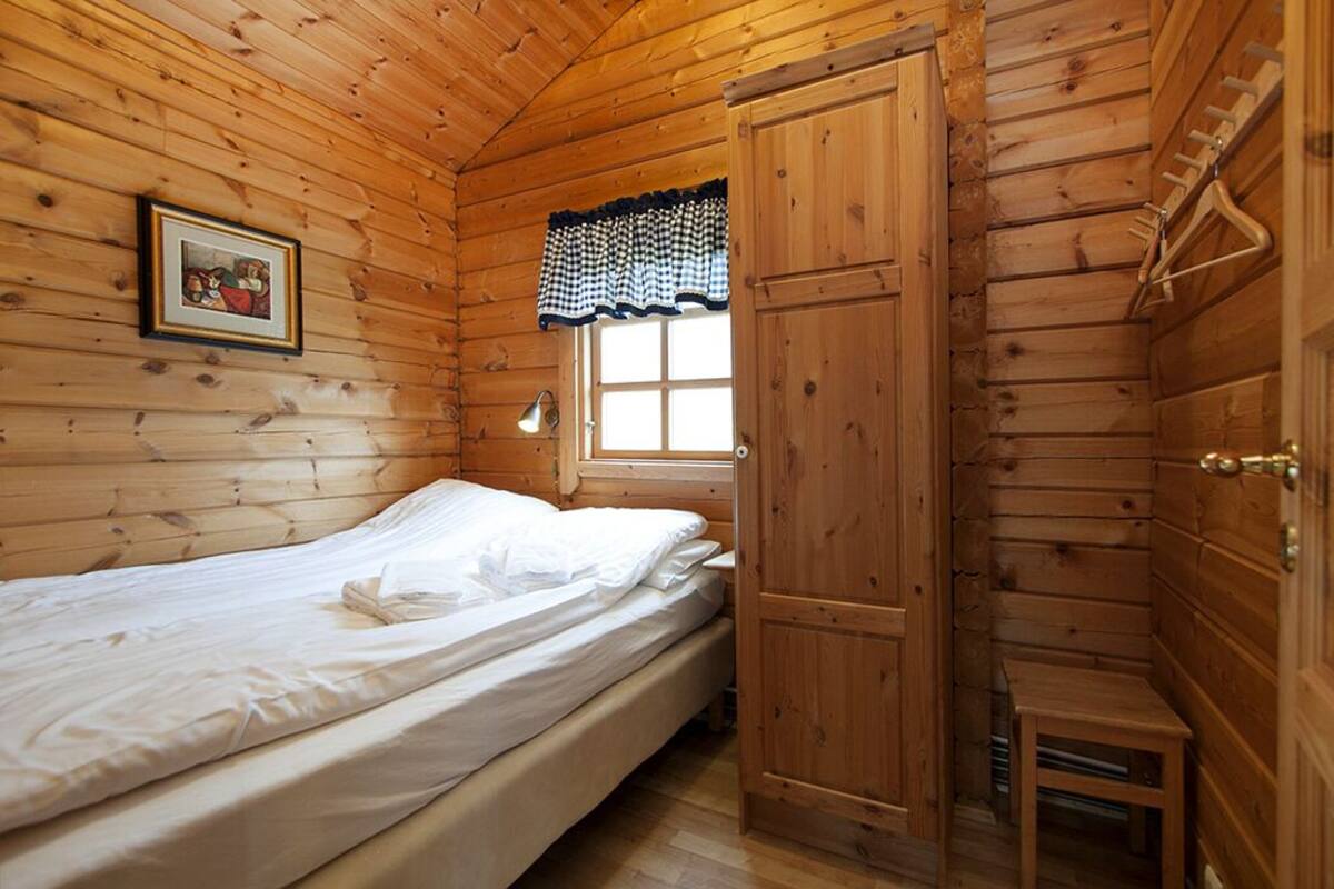 A wooden cabin room with a bed, a window with a checkered curtain, a door, and a small table.