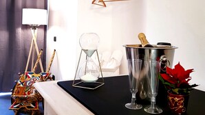 A hotel room with a black table, a metal bucket with a bottle, a glass, and a red cup. There is a white dresser with drawers, a colorful bag, and a white chair. - Sicilian Rooms Palermo (Palermo)