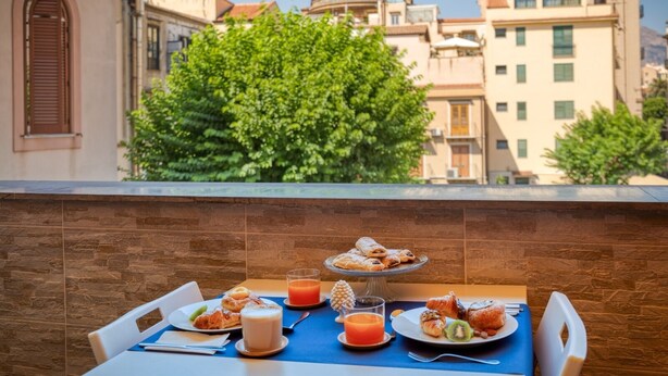 A table set with breakfast items including pastries, a glass of orange juice, and a cup of coffee, overlooking a cityscape with buildings and a tree. - Sicilian Rooms Palermo (Palermo)