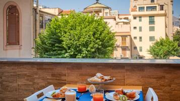 Una tavola imbandita per la colazione con pasticcini, un bicchiere di succo d'arancia e una tazza di caffè, con vista su un paesaggio urbano caratterizzato da edifici e alberi.
