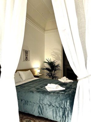 Una habitación de hotel con cama, mesita de noche, lámpara, planta y un cuadro en la pared.