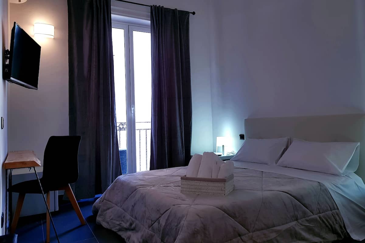 A hotel room with a bed, a TV, a desk with a chair, and a balcony with curtains.
