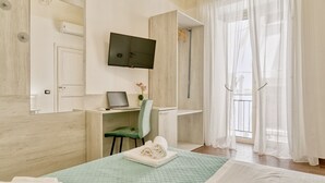 A hotel room with a bed, a desk with a laptop, a chair, a TV mounted on the wall, and a window with sheer curtains. - Sicilian Rooms Palermo (Palermo)