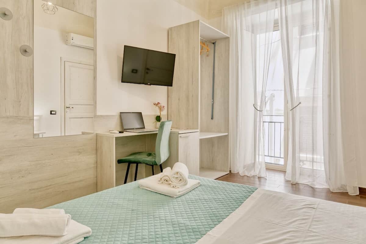 A hotel room with a bed, a desk with a laptop, a chair, a TV mounted on the wall, and a window with sheer curtains.