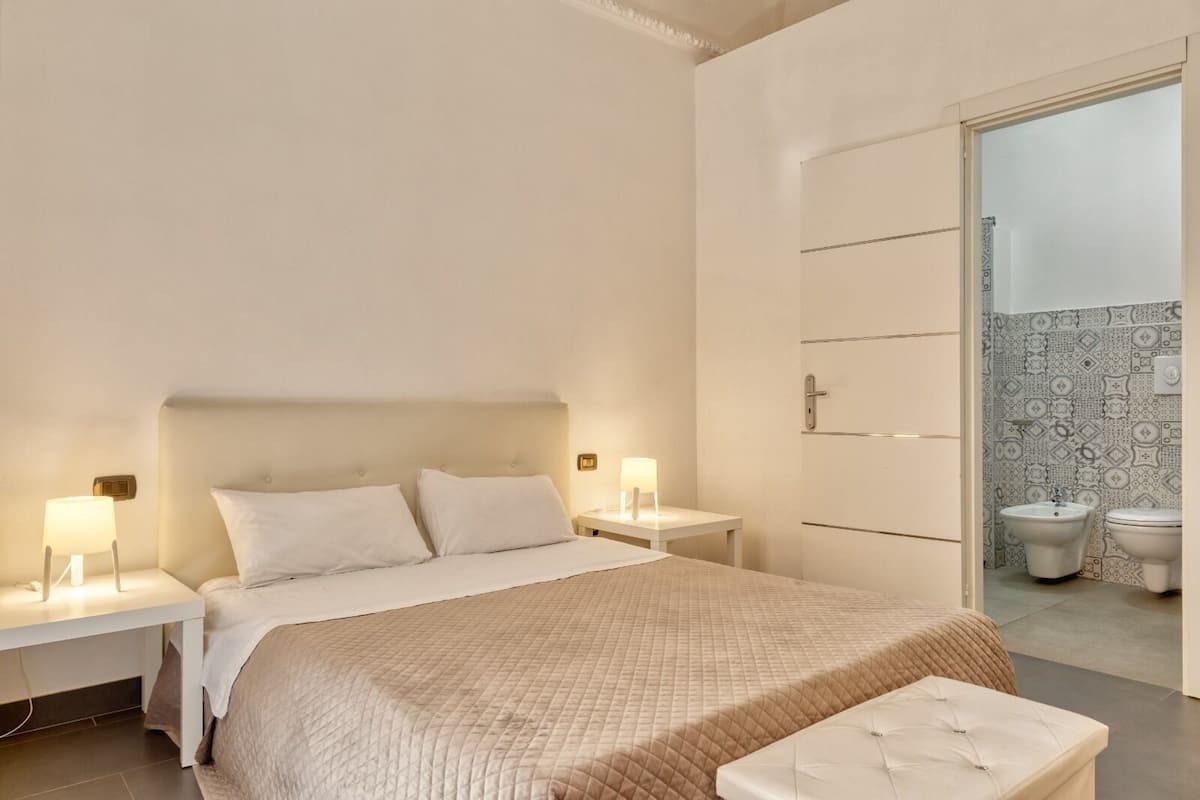 A hotel room with a bed, bedside tables with lamps, a bench, and a bathroom visible through an open door.