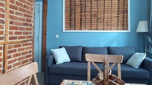 Une pièce avec un mur bleu, une fenêtre à cadre en bois avec des stores, un canapé de couleur foncée agrémenté de coussins, une chaise en bois et une petite table ornée d’un motif décoratif en carreaux.