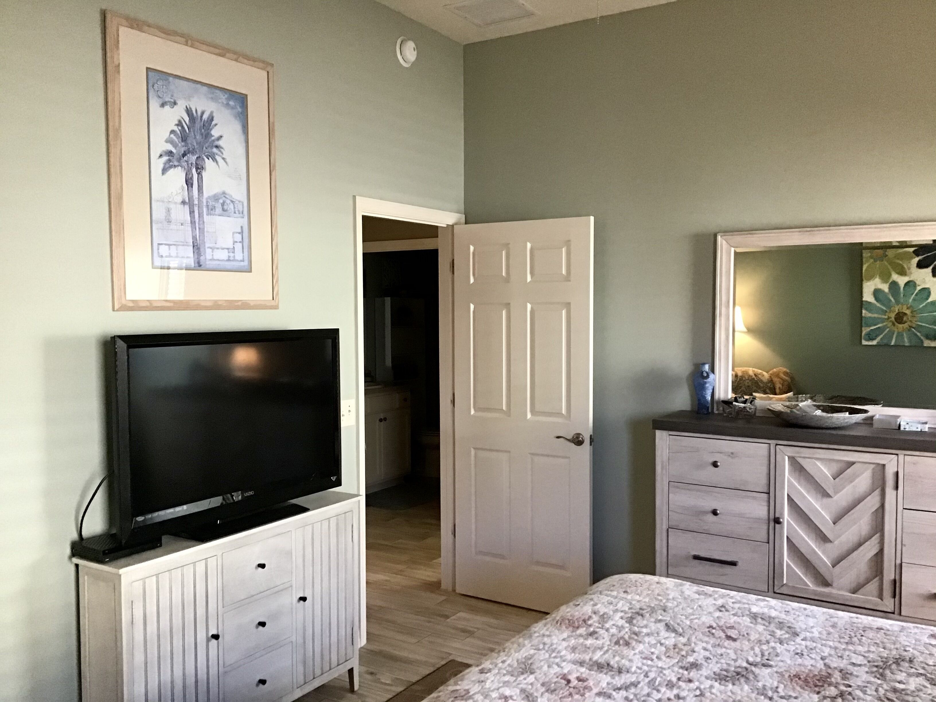 Een slaapkamer met een televisie op een kast, een deur naar een andere kamer, een dressoir met laden, en een bed met een bloemenprint sprei.