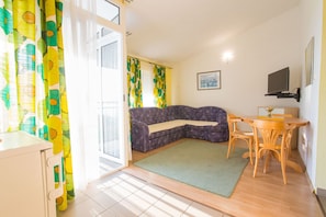 A cozy room with a sectional sofa, a small dining table with chairs, a TV mounted on the wall, and a window with curtains.