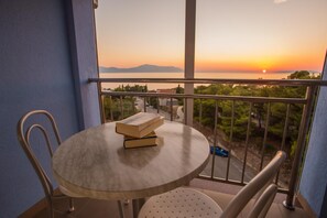 A balcony with a table and chairs, overlooking a sunset view with mountains and a body of water.